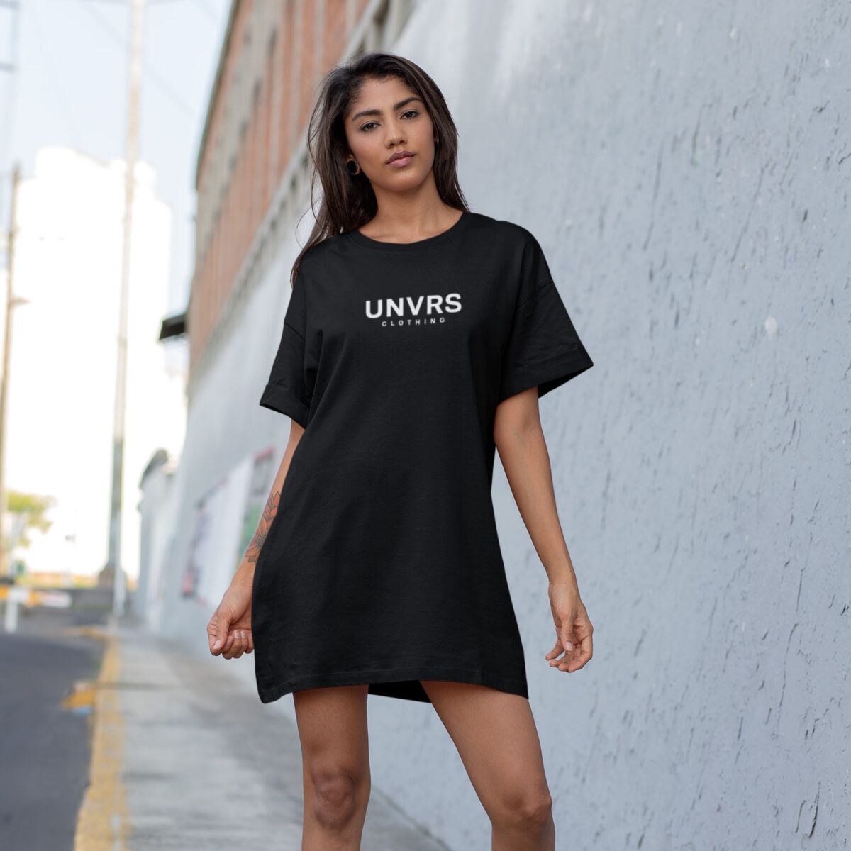 Female model wearing a UNVRS t-shirt dress, styled with a relaxed, oversized fit for a casual yet elevated look. The dress features minimalist branding, offering a versatile piece that can be worn alone or layered for a chic streetwear-inspired outfit. Ideal for summer days or night outings, embodying Ibiza’s bold and modern fashion vibe.